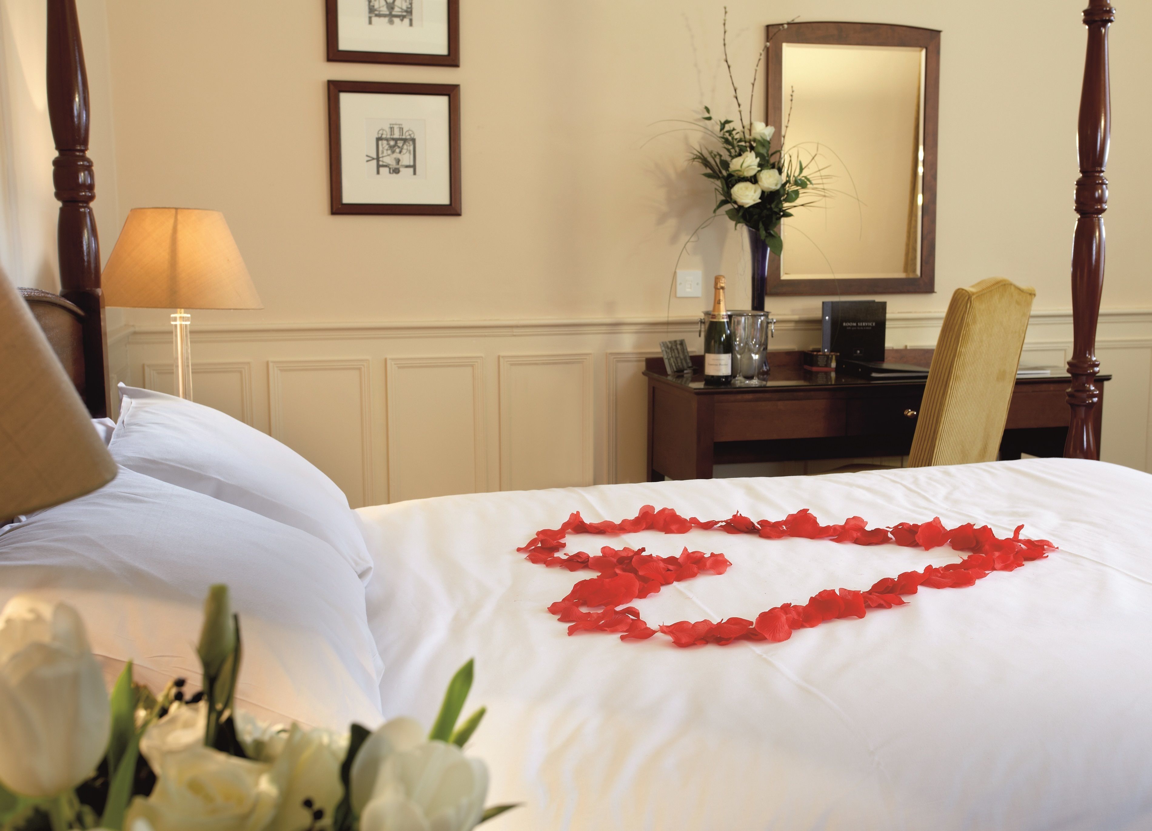 Una cama bien hecha con sábanas blancas y pétalos de rosa roja dispuestos en forma de corazón. Hay una lámpara, un jarrón con flores y un cuadro en la pared.