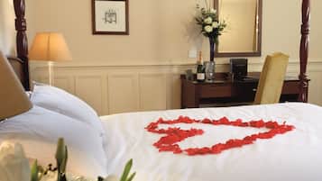 Una cama bien hecha con sábanas blancas y pétalos de rosa roja dispuestos en forma de corazón. Hay una lámpara, un jarrón con flores y un cuadro en la pared.