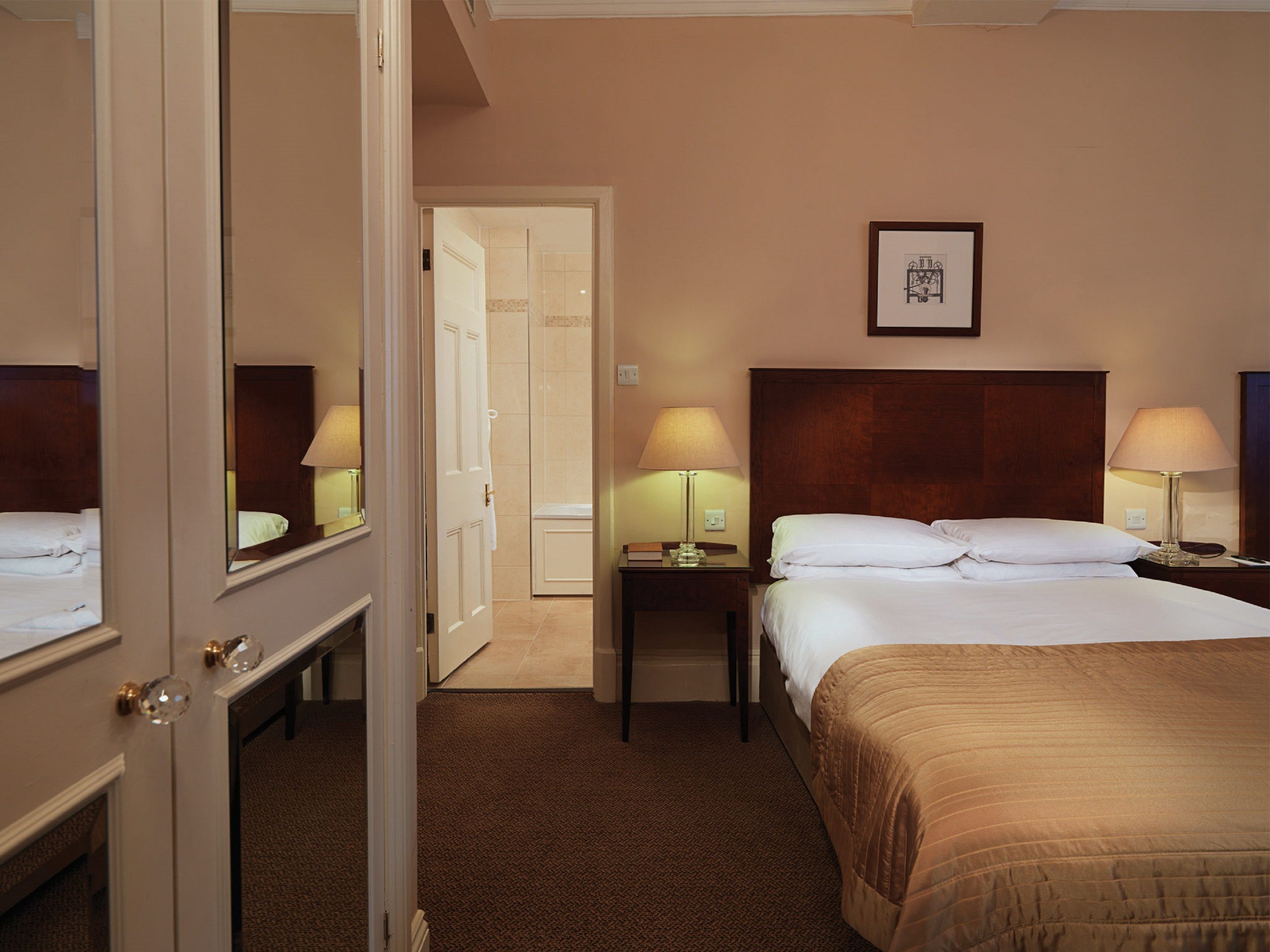 Een hotelkamer met een groot bed, twee nachtkastjes met lampen, een deur naar een andere kamer en een lijst aan de muur.