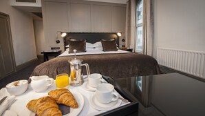 Habitación de hotel con una bandeja de desayuno ordenada sobre una mesita auxiliar, una cama con cabecera y una ventana con cortinas.