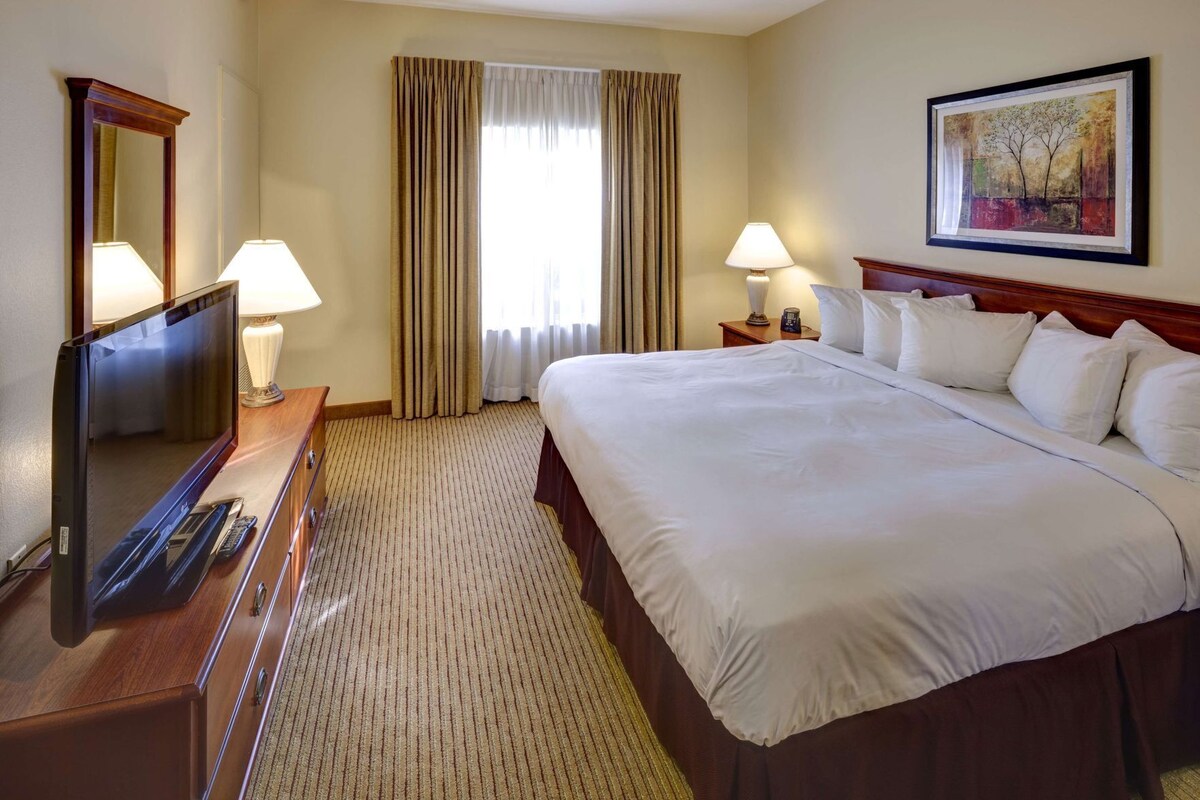 Habitación de hotel con una cama grande, un escritorio con televisor y control remoto, dos lámparas de noche, una ventana con cortinas y un cuadro en la pared.