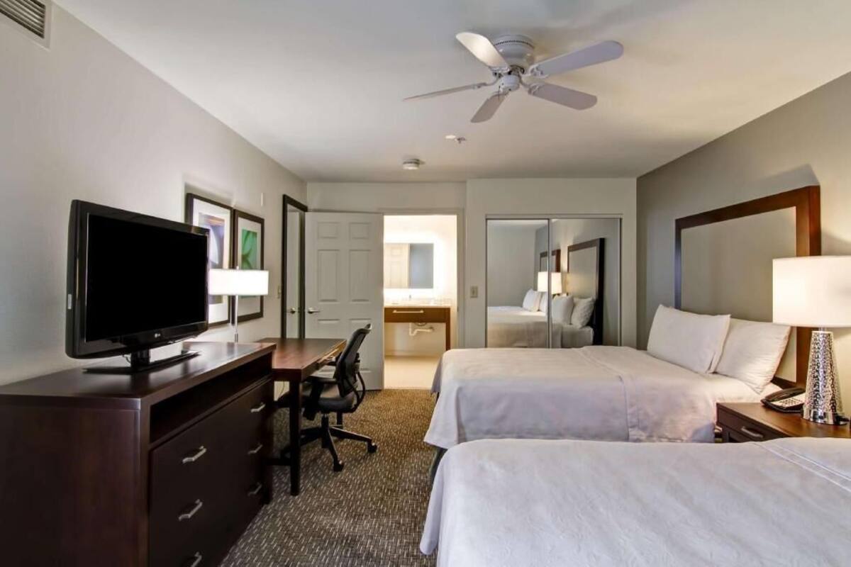 Een hotelkamer met een bed, een bureau met een computer, een televisie, een plafondventilator en een spiegel.