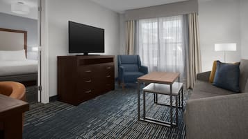 A hotel room with a bed, a TV, a desk, a chair, a sofa, and a window with curtains.