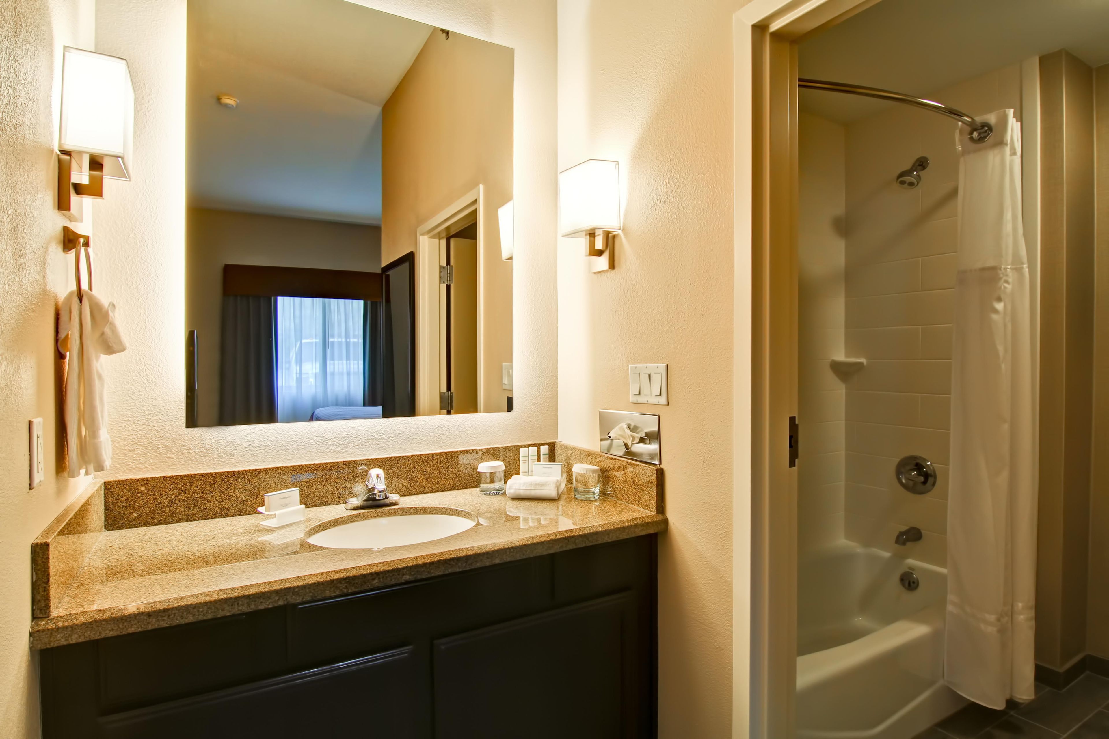 A hotel bathroom with a granite countertop, two sinks, a mirror, and a shower area.