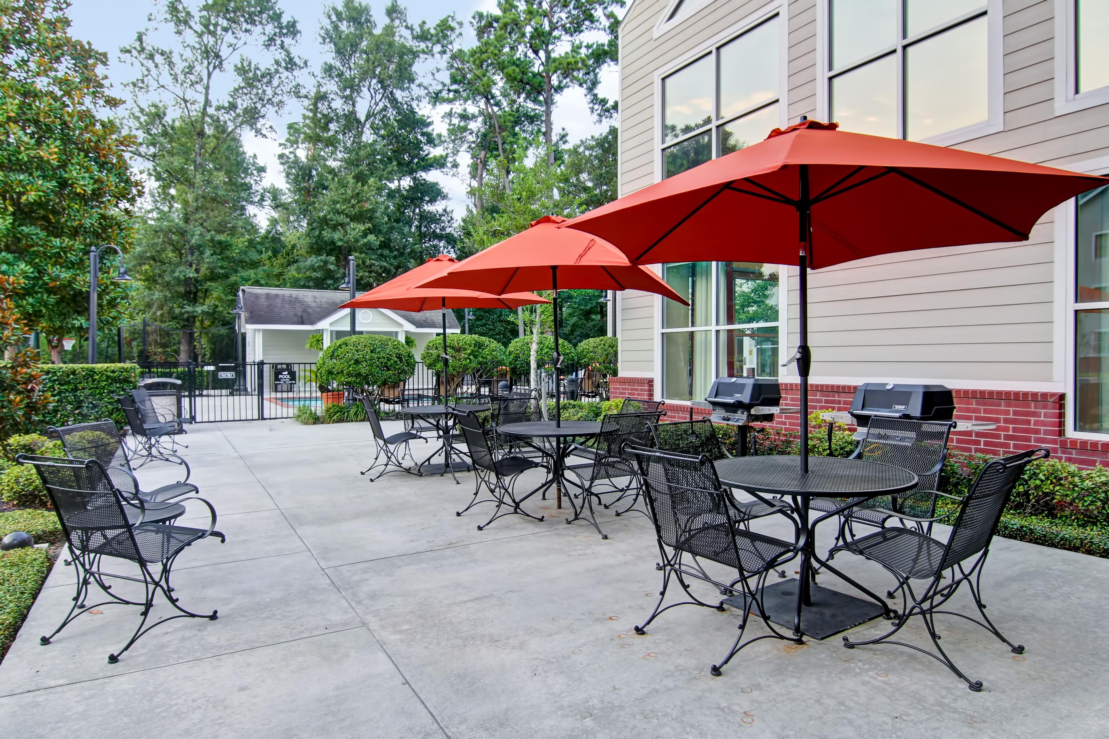 An outdoor patio with tables and chairs, red umbrellas, and a barbecue grill.