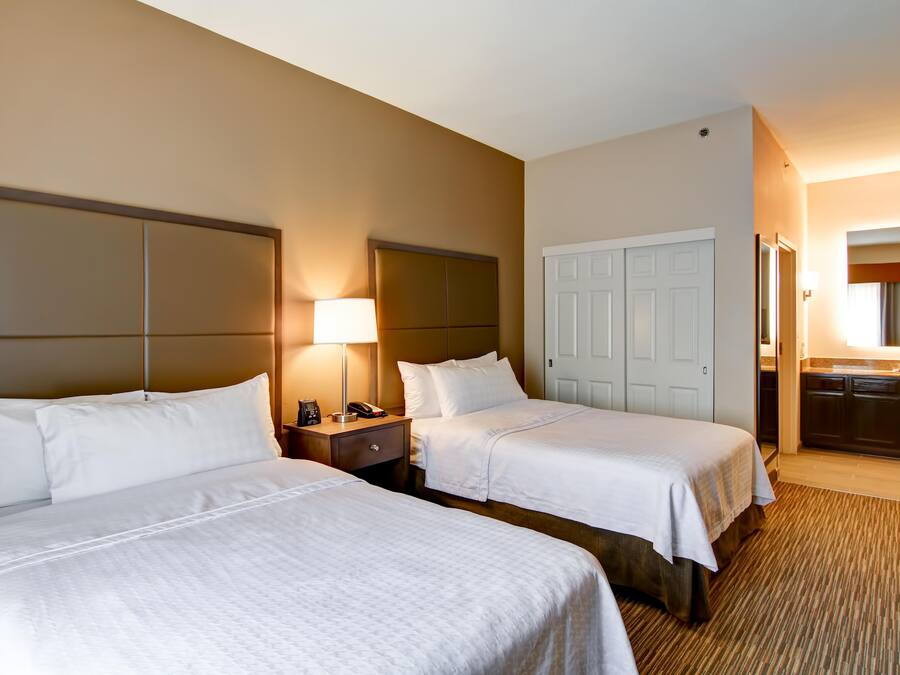A hotel room with two beds, a bedside table with a lamp, a wardrobe, and a bathroom visible through an open door.