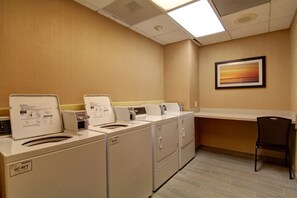 Une buanderie équipée de quatre machines à laver et de quatre sèche-linge, d’une chaise et d’un tableau accroché au mur.