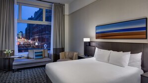 Habitación de hotel con una cama grande, una mesita de noche, una silla, un sofá y una ventana con vistas a la ciudad.