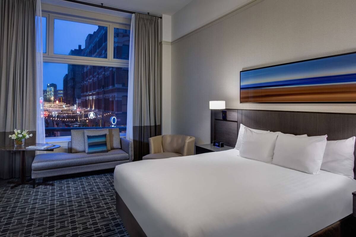 A hotel room with a large bed, a nightstand, a chair, a sofa, and a window with a city view.