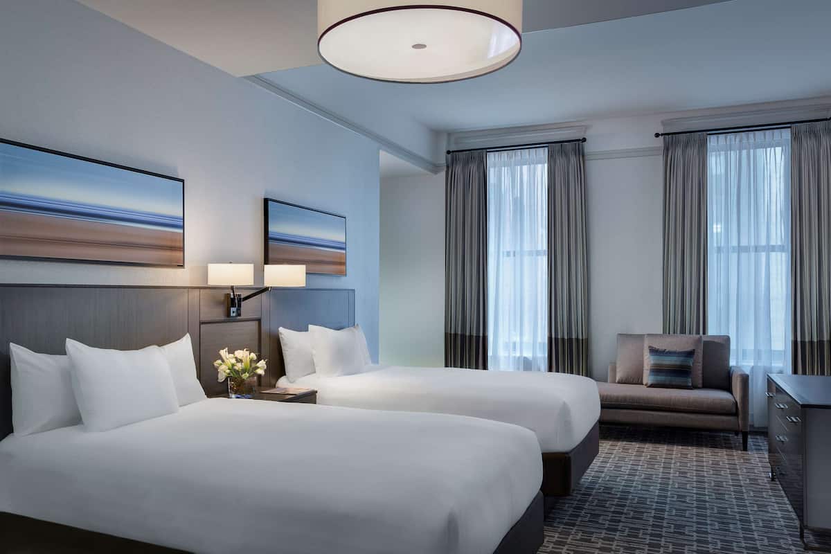 A hotel room with two beds, a desk, a chair, and a large window with curtains.