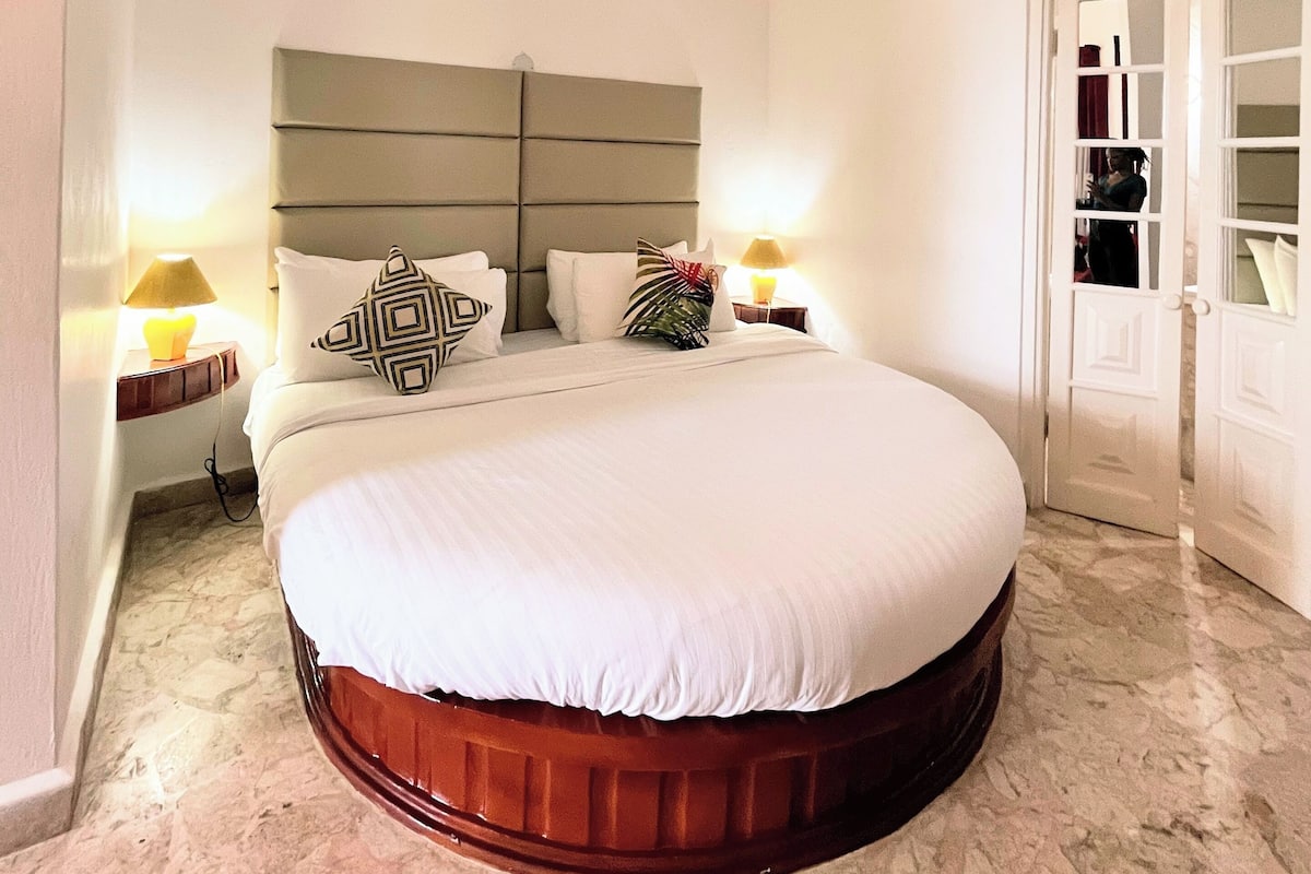 Un grand lit rond avec une base en bois, deux tables de chevet avec des lampes, une tête de lit blanche et une porte avec des panneaux de verre.