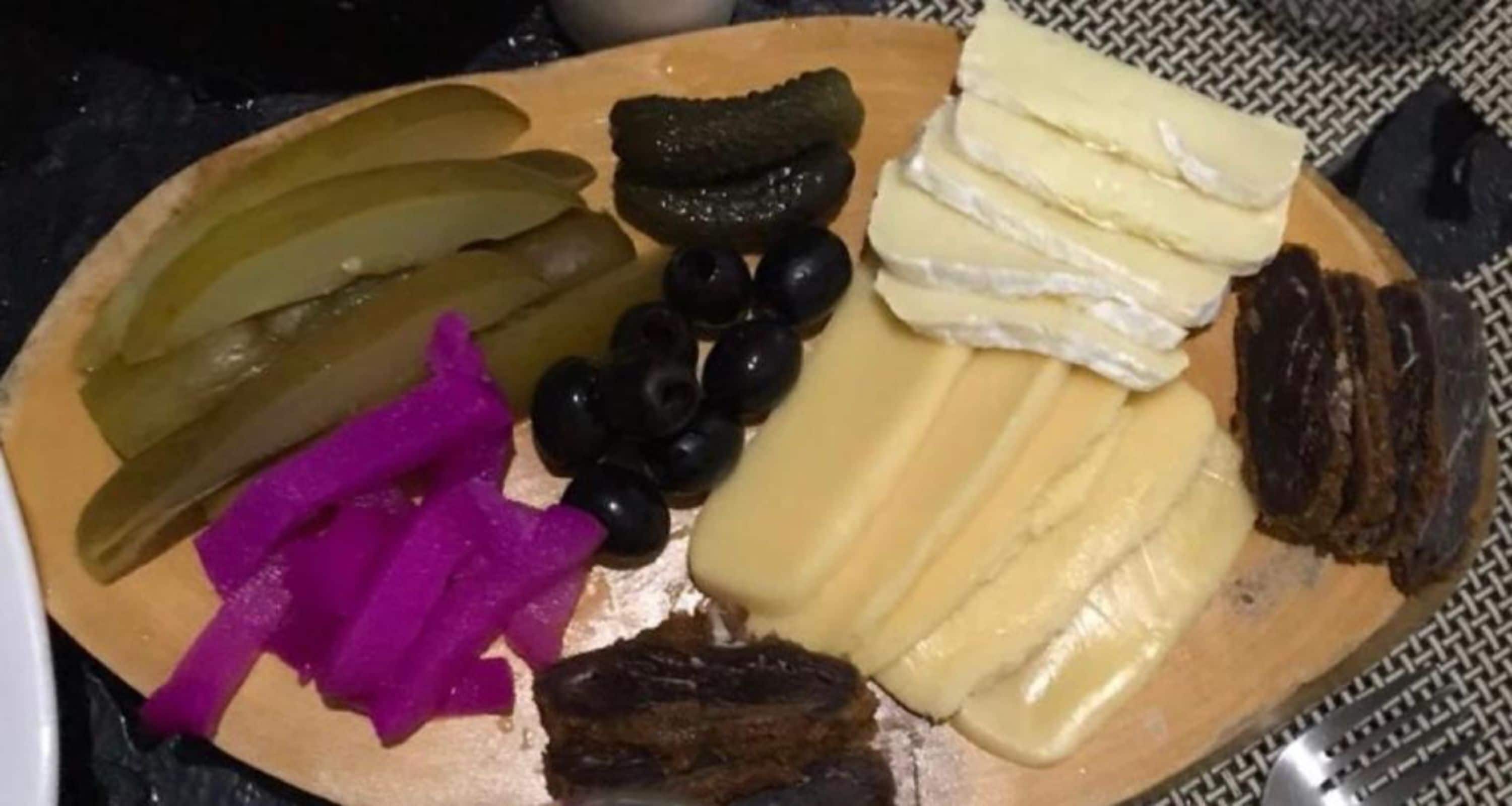 Una tabla de madera con diversos alimentos, como pepinillos, aceitunas, queso y un trozo de carne.
