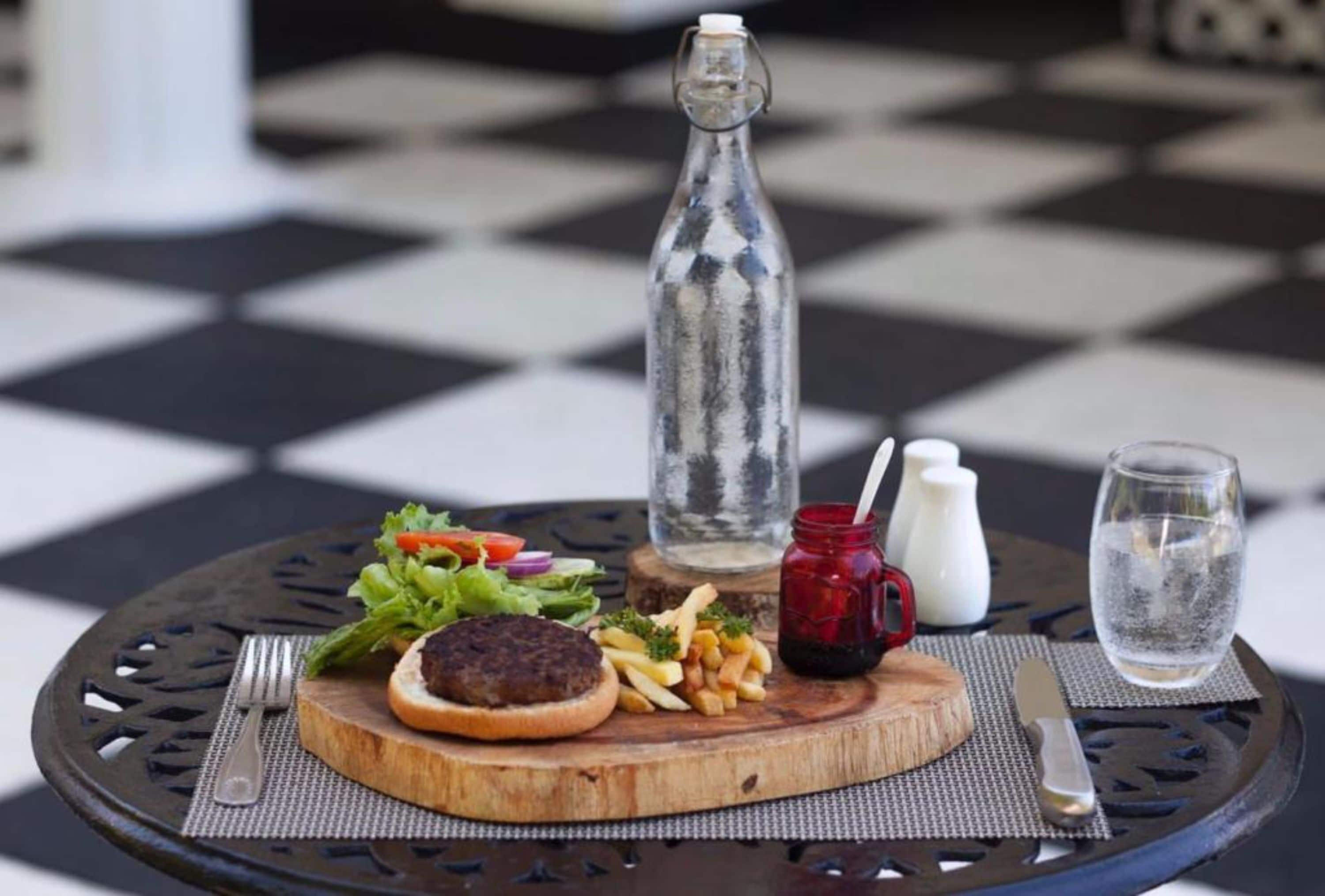 A meal with a burger, fries, and salad on a wooden board, a glass bottle, a red mug, a glass of water, and a fork on a round table with a black and white checkered floor.
