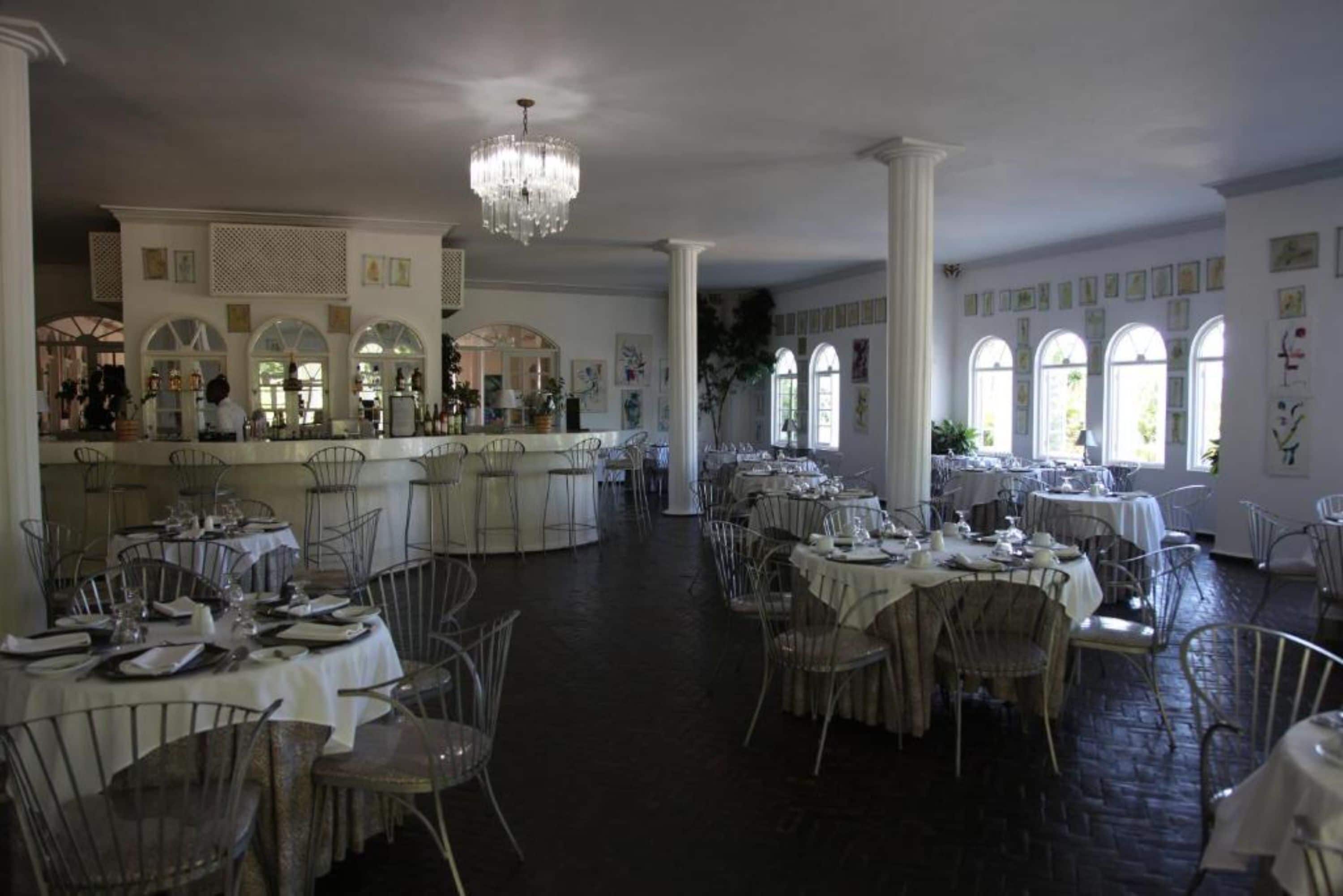 Un vaste espace dédié à la salle à manger, avec des tables et des chaises rondes, un coin bar agrémenté d’un lustre, et de grandes fenêtres surmontées d’arcs.