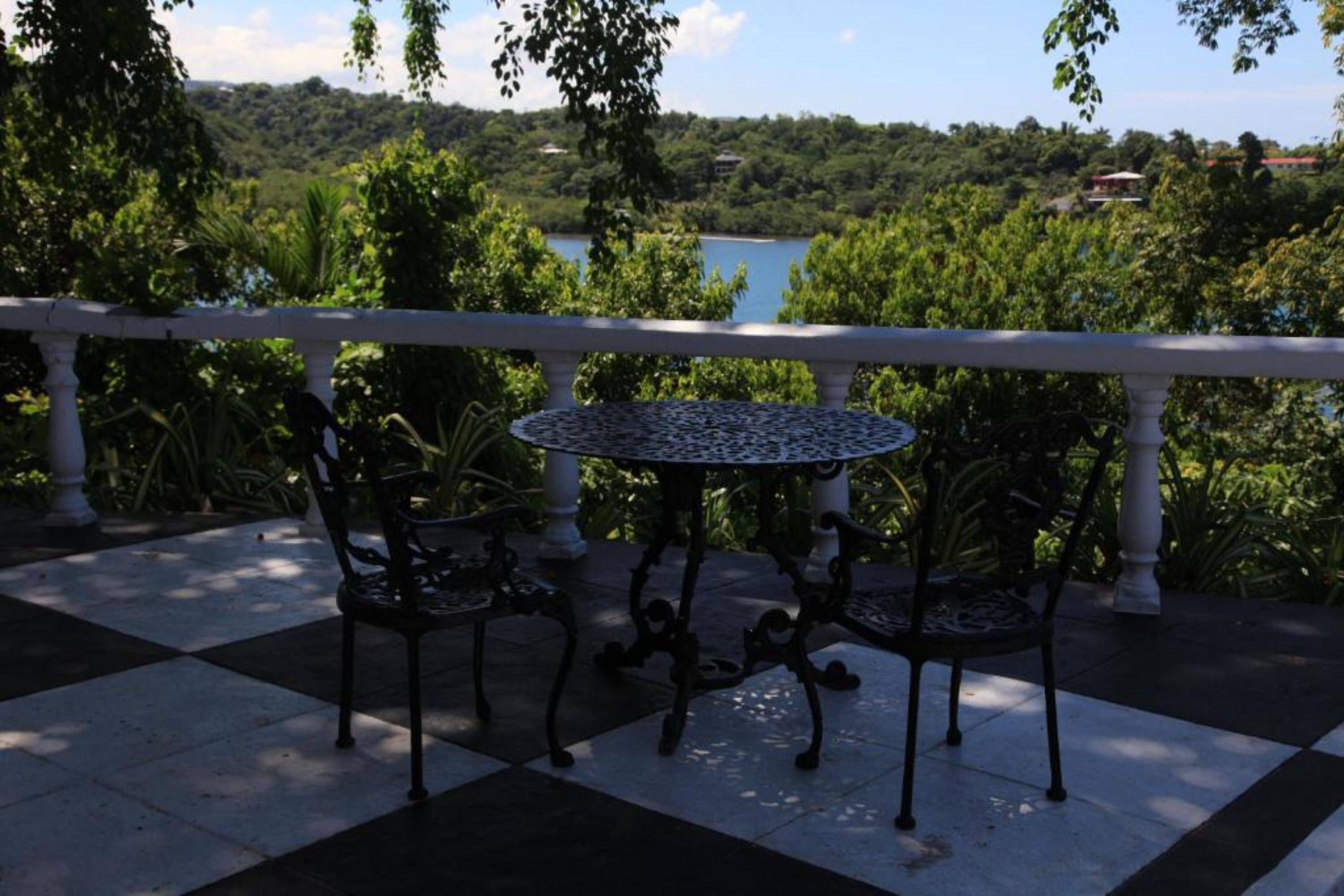 A terrace with a checkered floor, two chairs, a round table, and a white balustrade overlooking a lake and greenery.