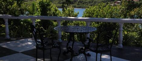 A terrace with a checkered floor, two chairs, a round table, and a white balustrade overlooking a lake and greenery.