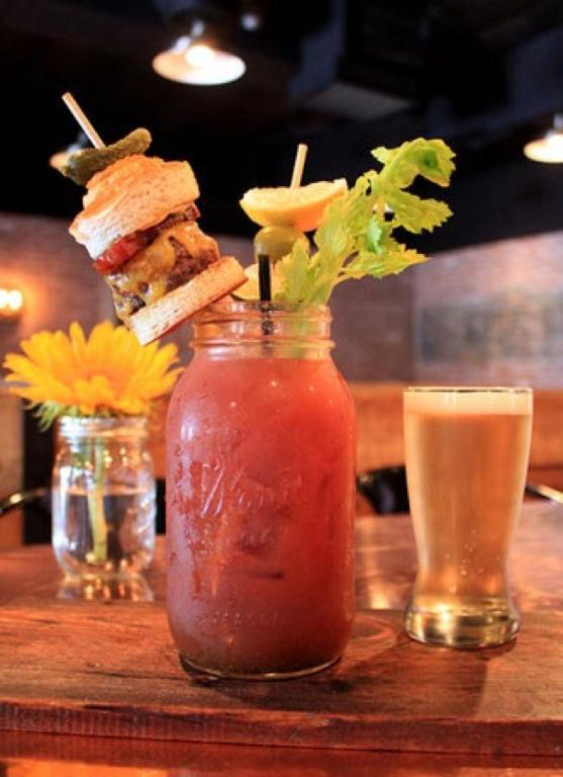 A glass of Bloody Mary with celery and a sandwich on a wooden table.