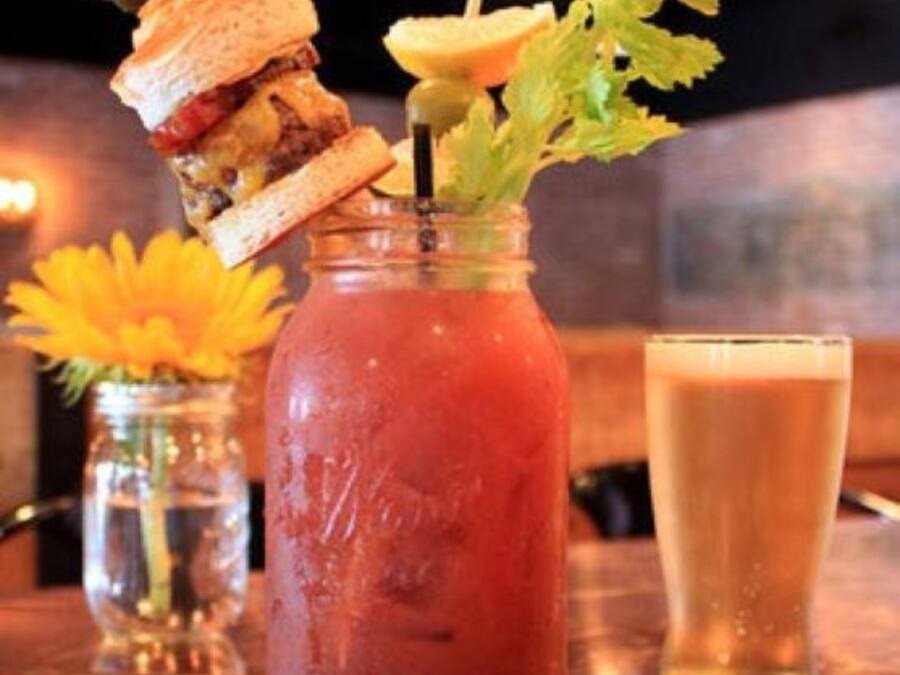 A glass of Bloody Mary with celery and a sandwich on a wooden table.