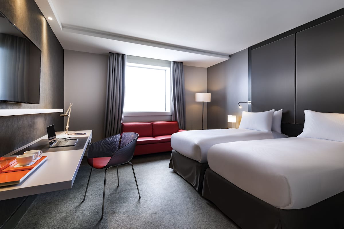 A hotel room with a desk, two beds, a red sofa, a TV, and a window with curtains.