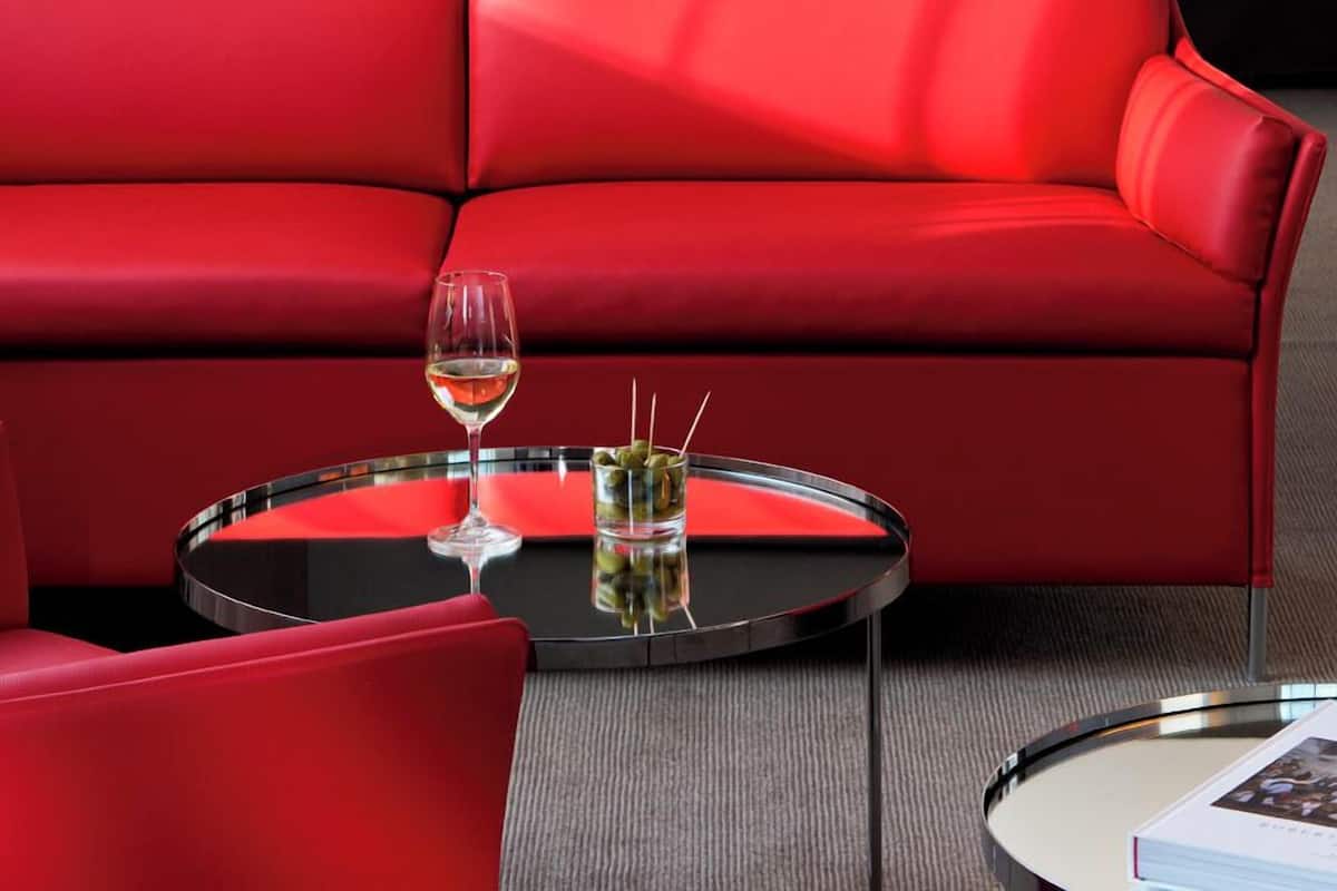 A modern hotel room with a red sofa, a glass coffee table with a wine glass, and a bed with white linens.