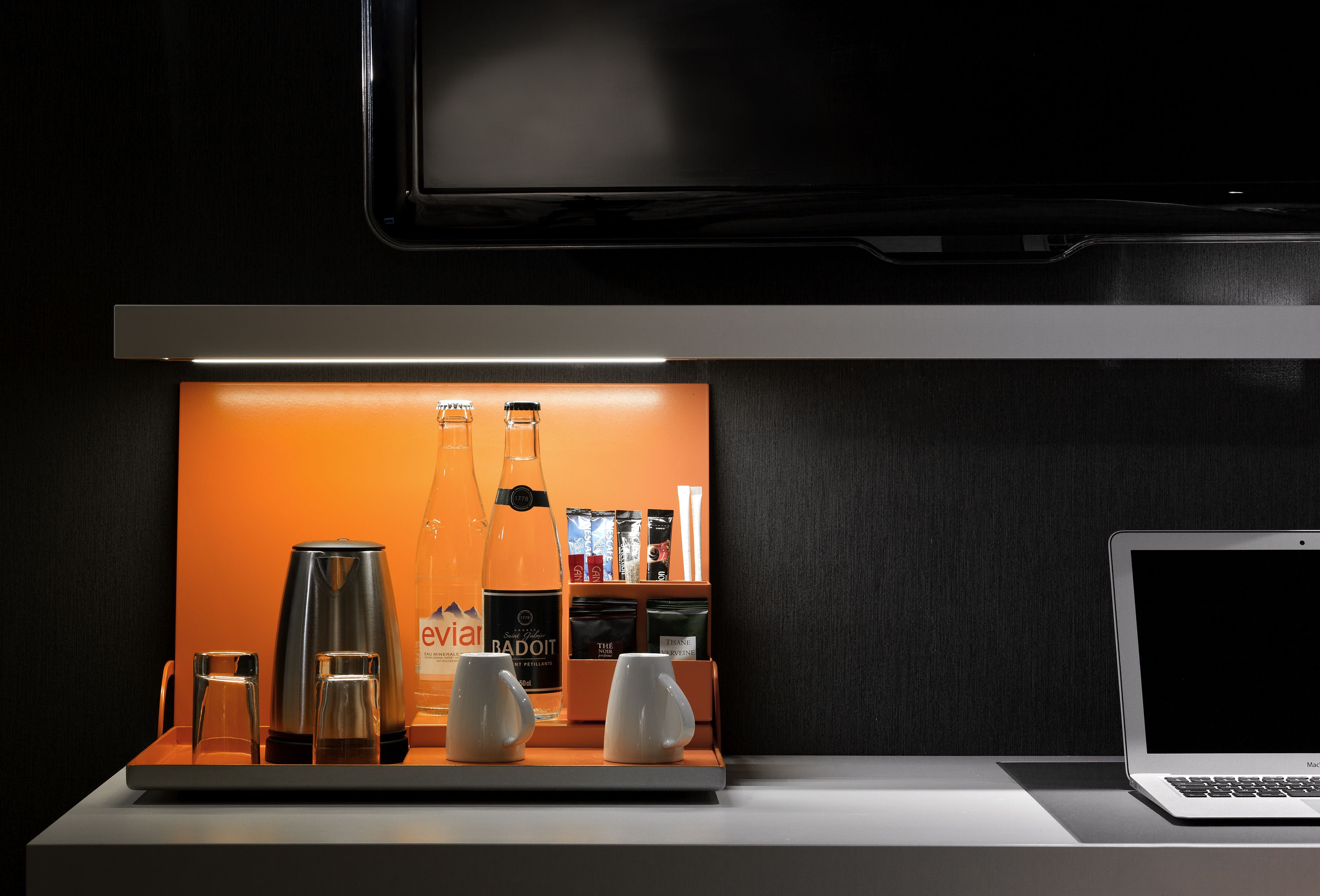 Een moderne hotelkamer met een strakke, donkere houten afwerking, een flatscreen televisie aan de muur en een minimalistische koffiehoek met een koffiezetapparaat, twee glazen en een fles Badoit.