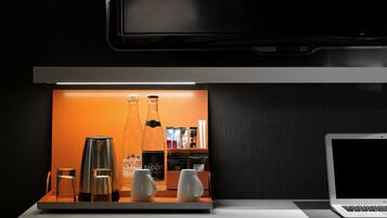 Een moderne hotelkamer met een strakke, donkere houten afwerking, een flatscreen televisie aan de muur en een minimalistische koffiehoek met een koffiezetapparaat, twee glazen en een fles Badoit.