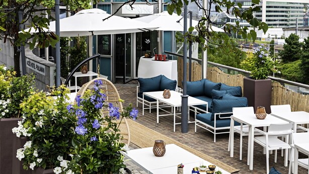 Un patio al aire libre con mesas blancas y asientos acolchados en azul, rodeado de plantas y flores en macetas.