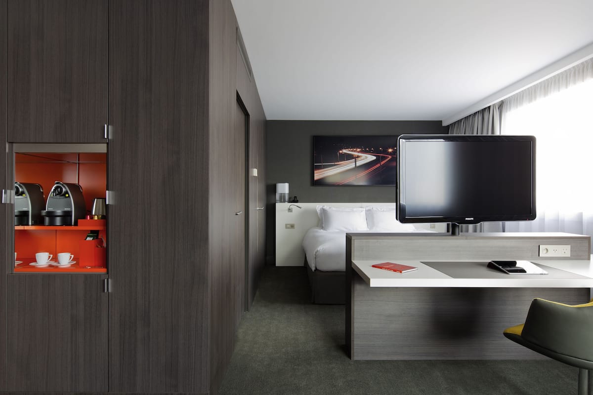 A modern hotel room with a flat-screen TV, a desk with a chair, a bed, and a minibar.