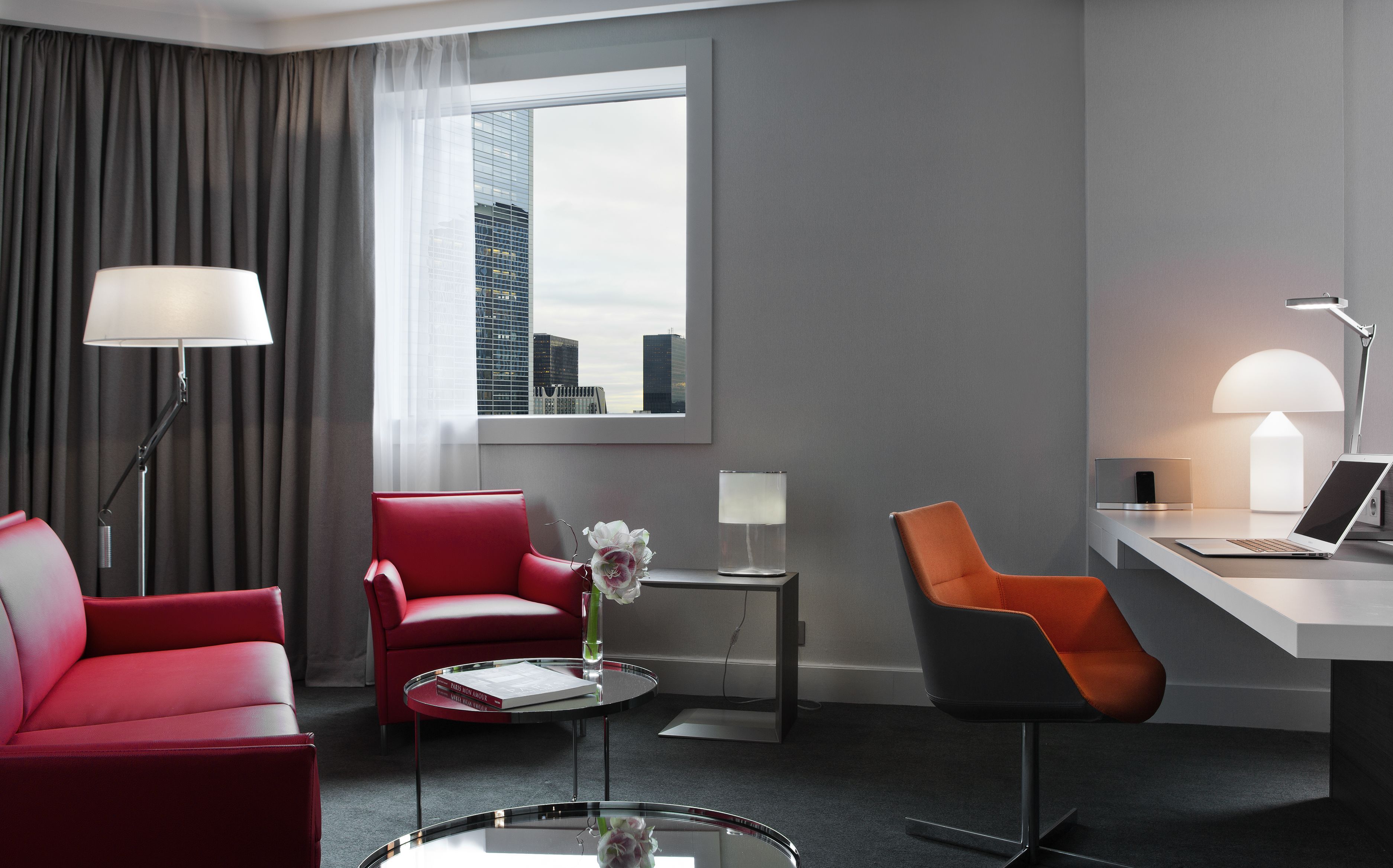 A modern hotel room with a red armchair, a grey armchair, a desk with a laptop, and a large window with a city view.