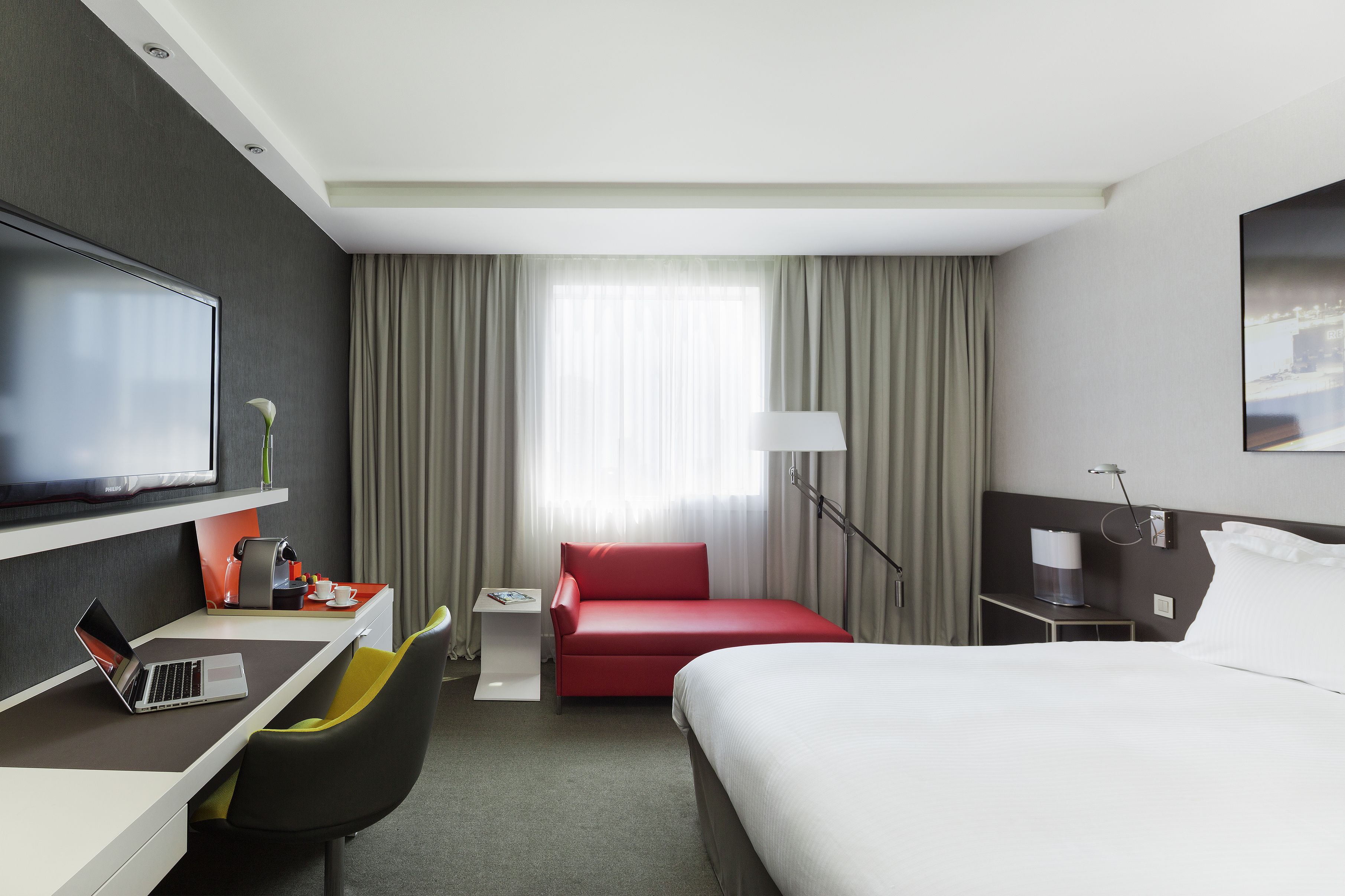 A hotel room with a bed, a desk with a laptop, a red sofa, a TV, and a window with curtains.