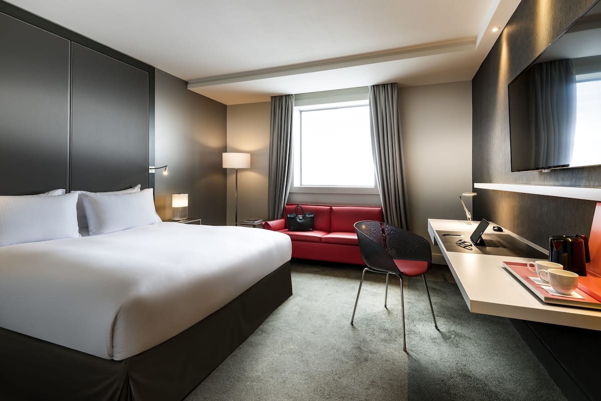 A modern hotel room with a large bed, a desk, a red sofa, and a TV mounted on the wall.