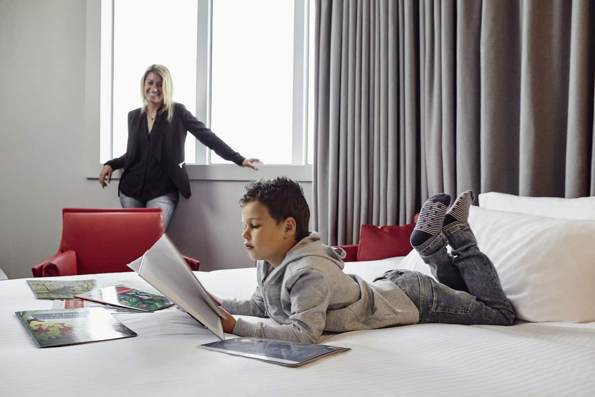 A hotel room with a bed, a laptop, and a tablet on the bed. There is a red chair and a window with curtains.