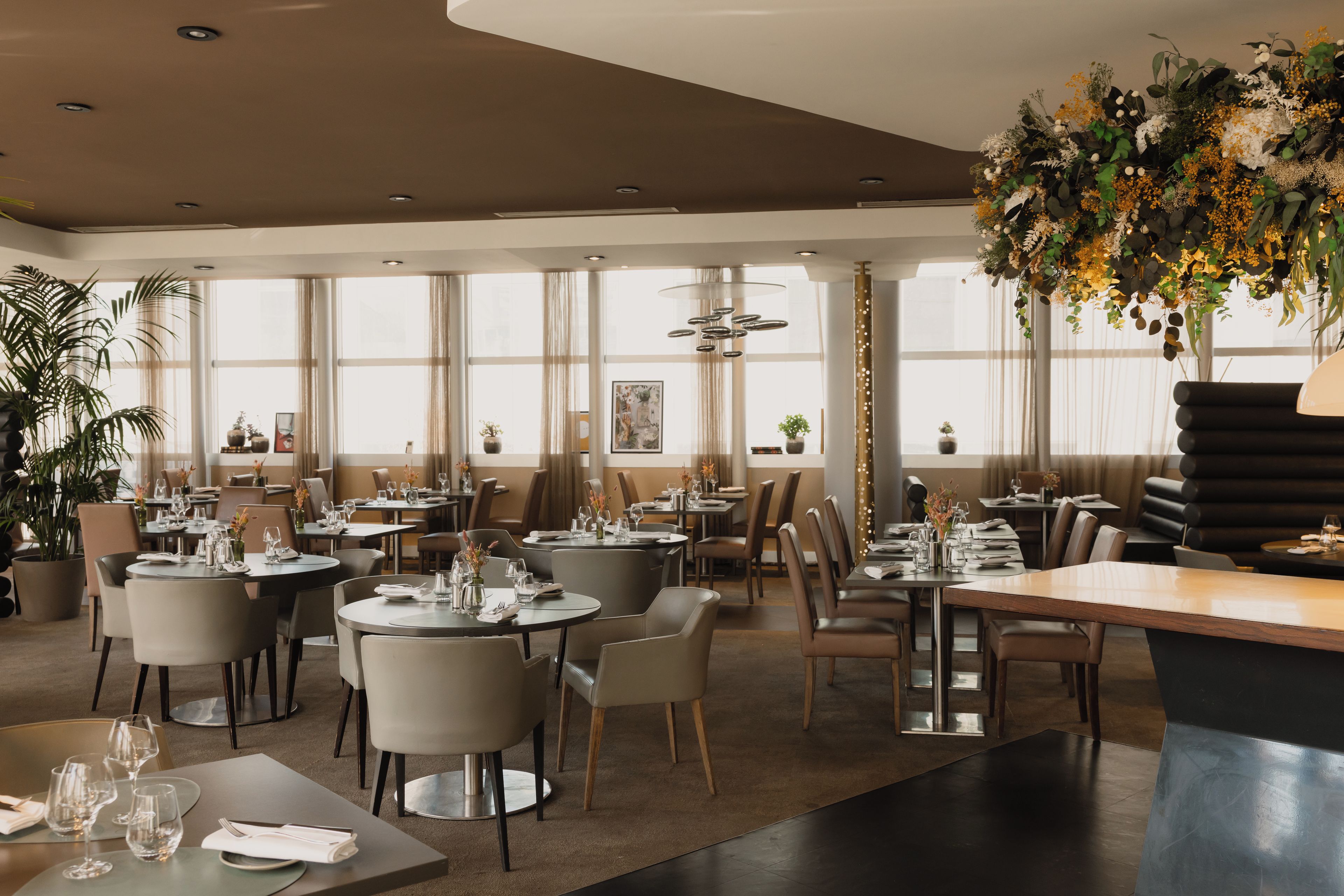 A modern restaurant interior with round tables, chairs, and a large floral centerpiece.