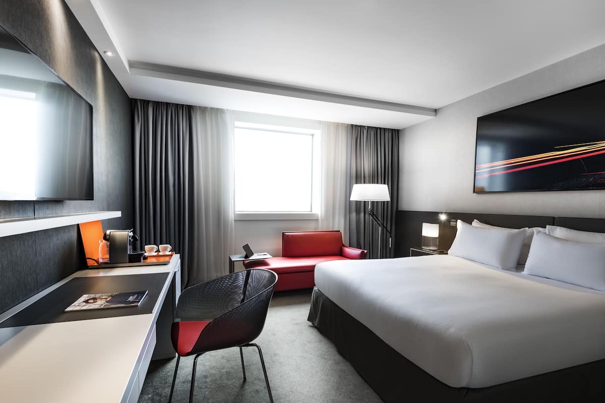 A modern hotel room with a large bed, a desk with a laptop, a red sofa, and a large window with curtains.