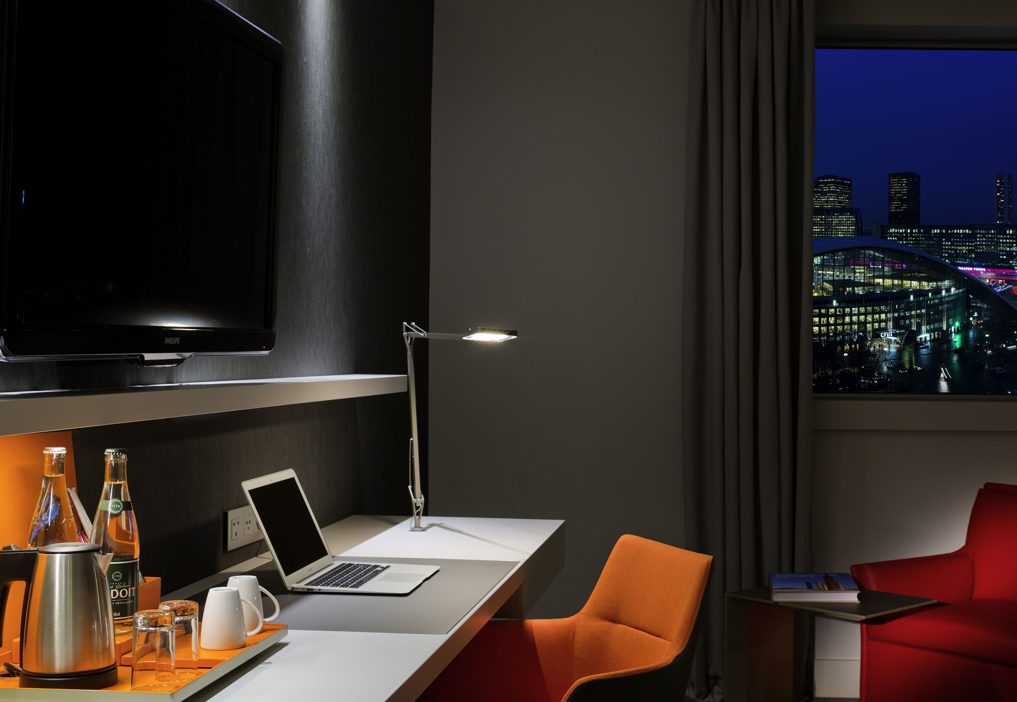 A modern hotel room with a desk, laptop, and a large TV. There are two chairs, a red one and an orange one. A cityscape is visible through the window.