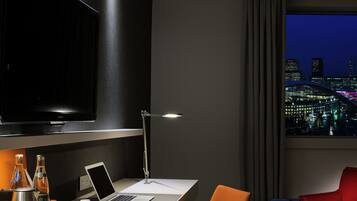 A modern hotel room with a desk, laptop, and a large TV. There are two chairs, a red one and an orange one. A cityscape is visible through the window.