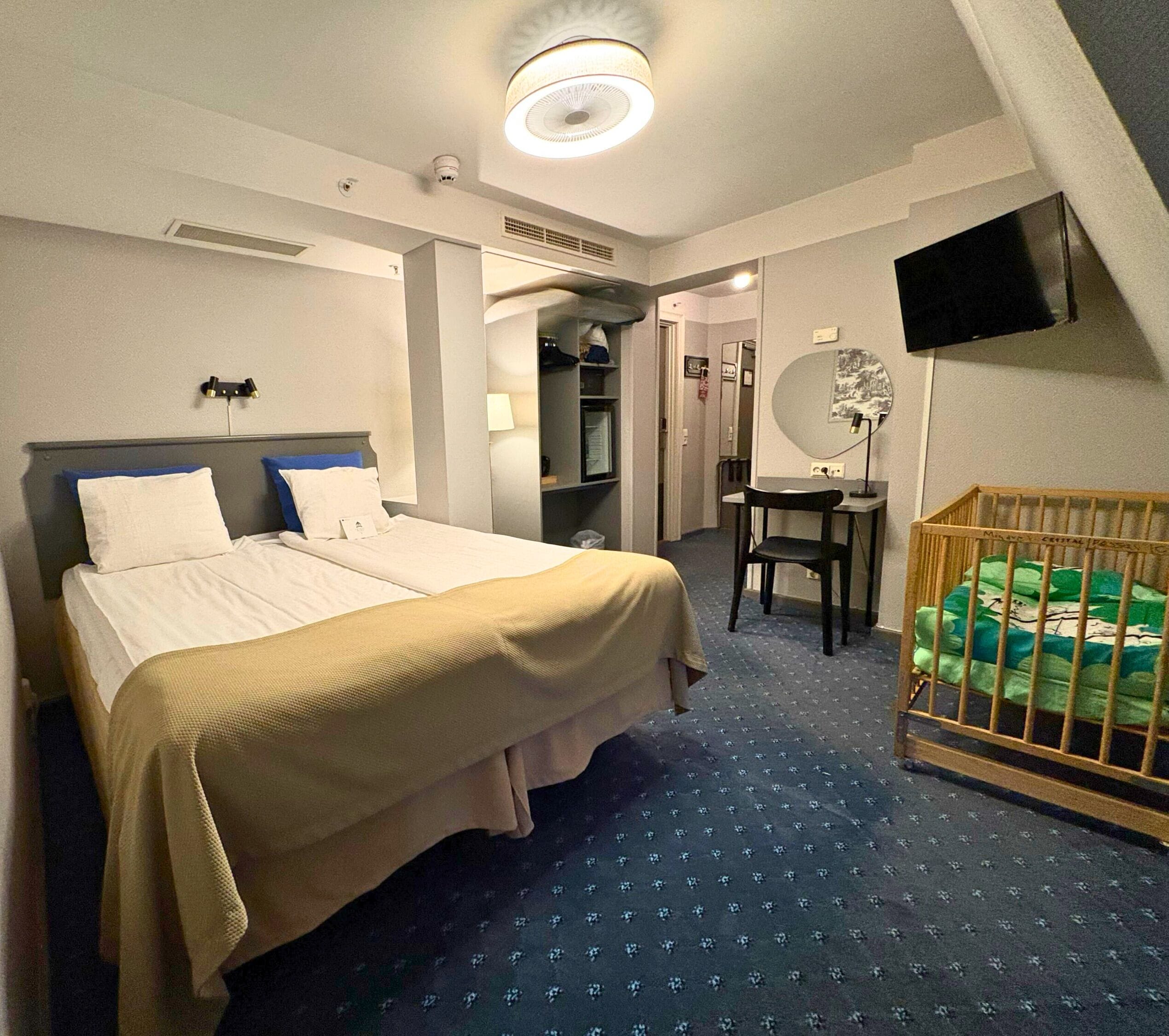 A hotel room with a bed, a desk, a chair, a crib, a television, and a mirror.