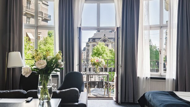 A hotel room with a bed, a desk with a lamp, a chair, a vase with flowers, and a view of the city through large windows.