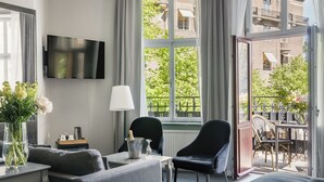 A modern living room with a sofa, a table with a vase of flowers, a TV mounted on the wall, and a window with a view of buildings and greenery.