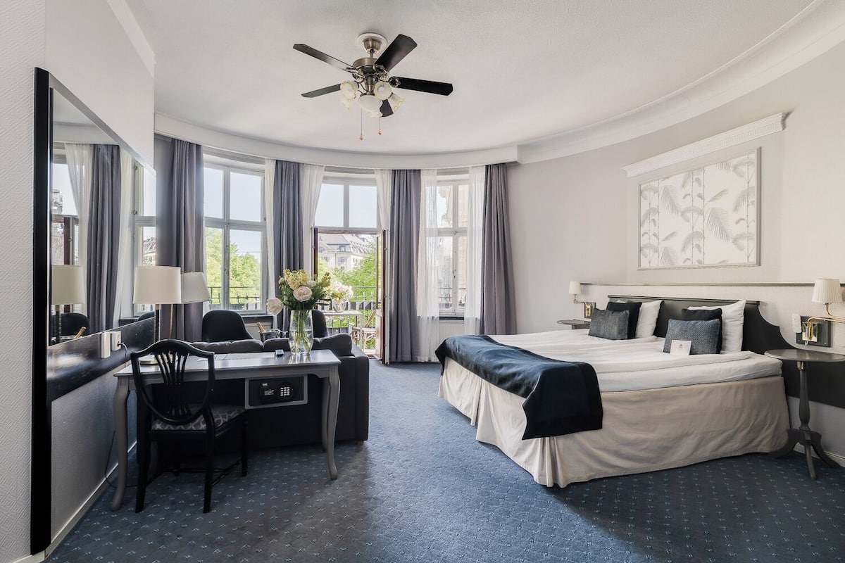 Ett hotellrum med en säng, ett skrivbord, en stol och utsikt över staden genom stora fönster.
