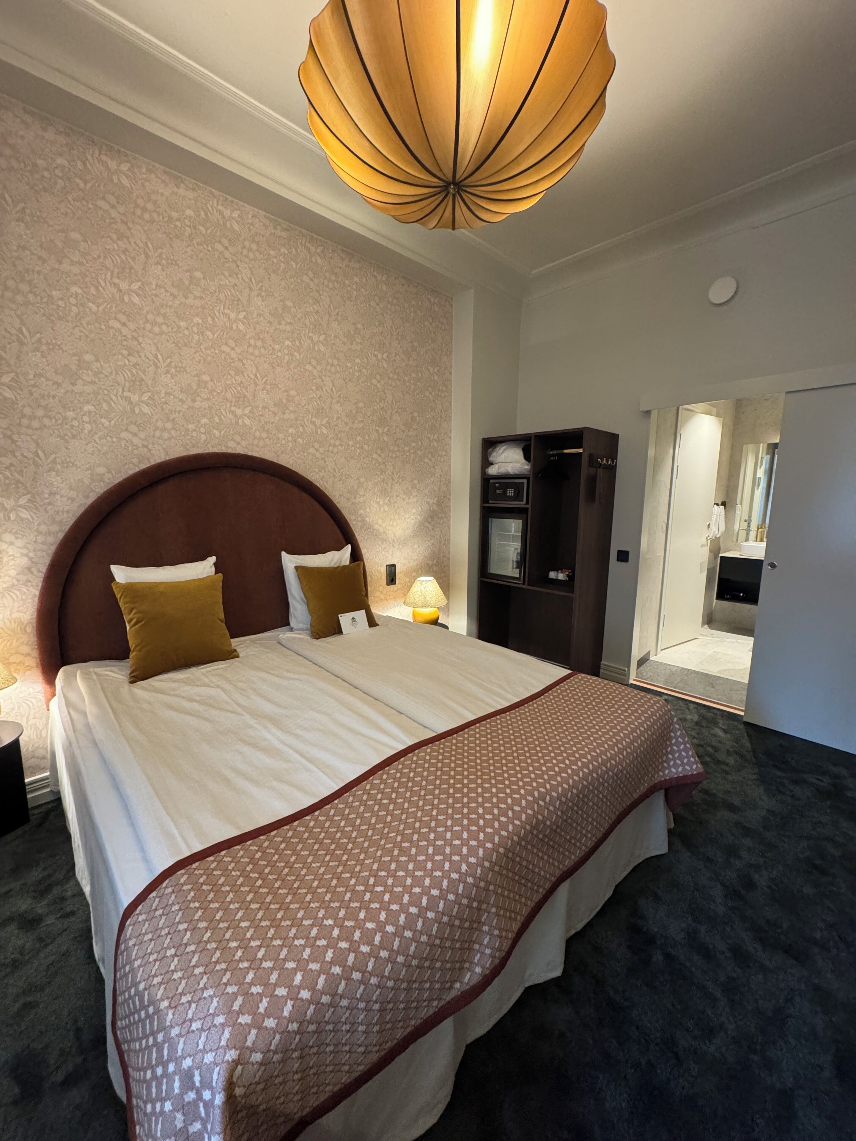 Ett hotellrum med en stor säng, en sänggavel i trä, ett mönstrat överkast, en modern kristallkrona, en minibar och ett badrum som syns genom en öppen dörr.
