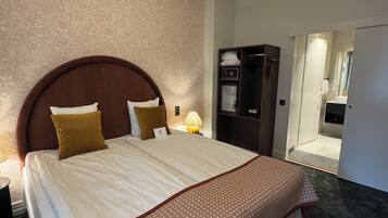 Ett hotellrum med en stor säng, en sänggavel i trä, ett mönstrat överkast, en modern kristallkrona, en minibar och ett badrum som syns genom en öppen dörr.