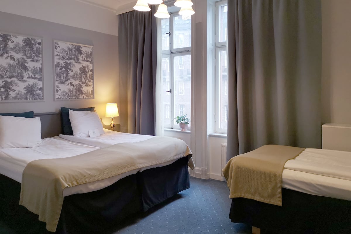 Ett hotellrum med två sängar, ett fönster med gardiner, en lampa och konst på väggen.