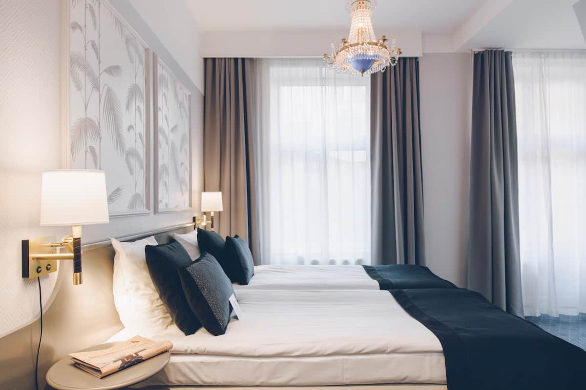 Ett hotellrum med en stor säng, två sängbord, en kristallkrona och ett stort fönster med gardiner.
