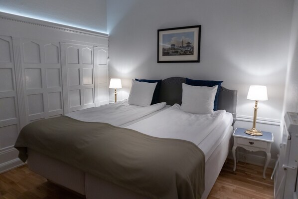 A neatly made double bed with white linens and a brown throw blanket, flanked by two bedside tables with lamps, in a room with white paneled walls and a framed picture above the bed. - Crystal Plaza Hotel (Stockholm)