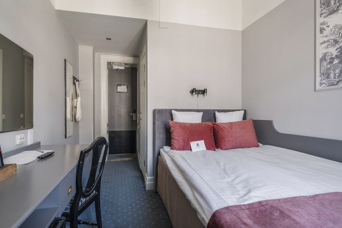 Ett hotellrum med en säng, ett skrivbord, en stol, en spegel och en dörr som leder till ett badrum.