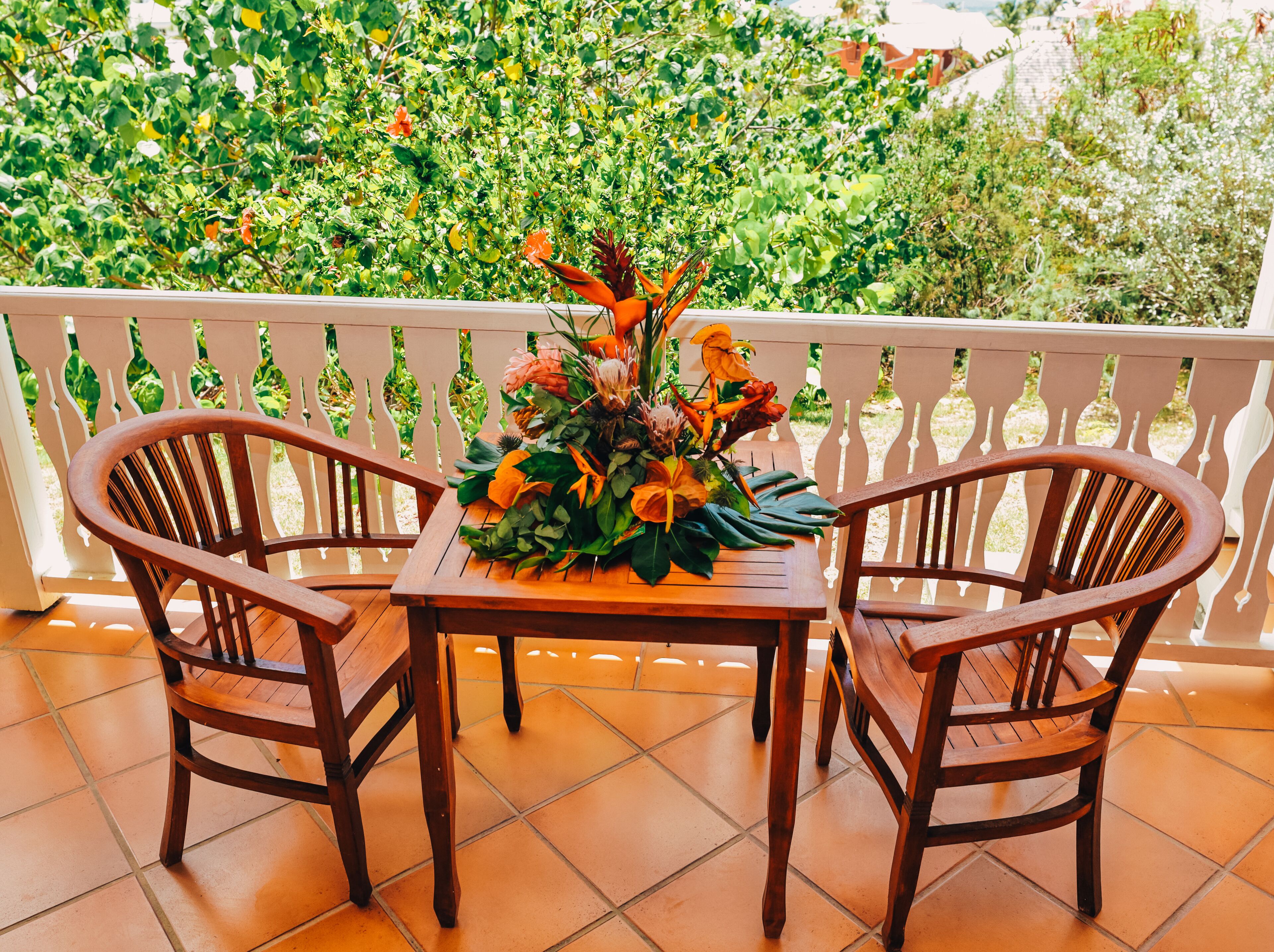 Uma mesa de madeira com cadeiras numa varanda com guarda de proteção branca, rodeada por vegetação exuberante.