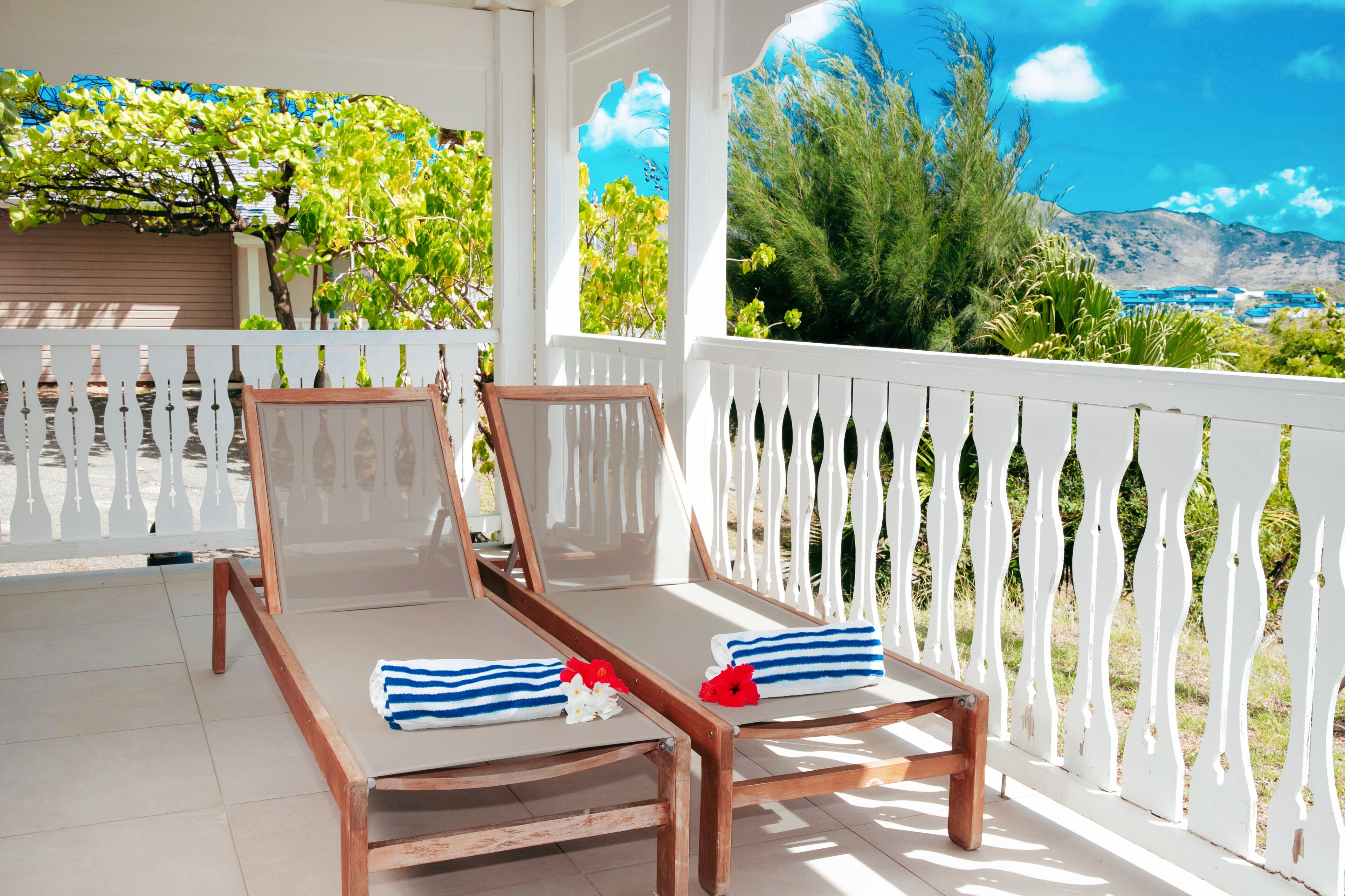 Two wooden lounge chairs with white cushions and blue-striped towels, a white picket fence, and a view of a mountainous landscape.
