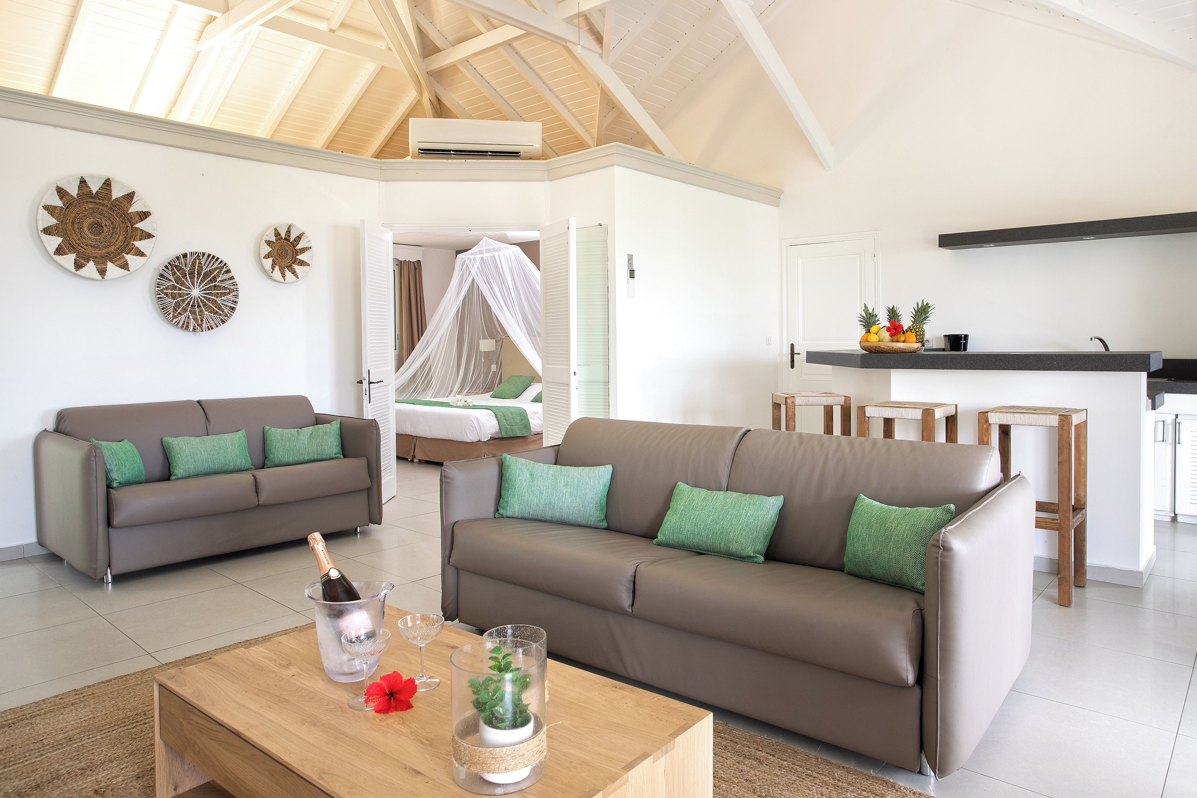 A modern living room with a high ceiling, a sofa set, a coffee table with a bottle and glass, and a kitchen area in the background.