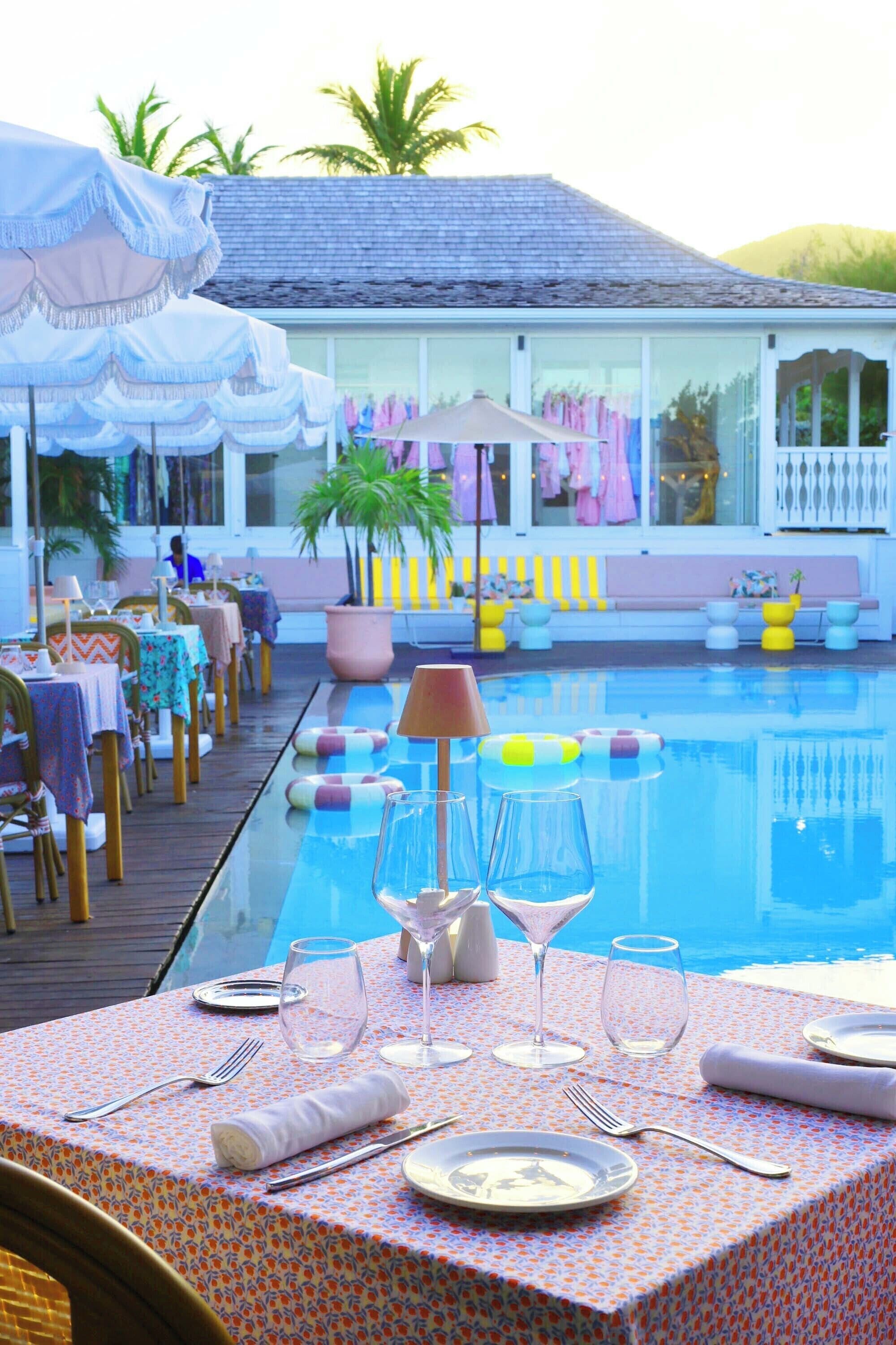 A poolside dining area with tables set for two, a pool, and a seating area with umbrellas and chairs.